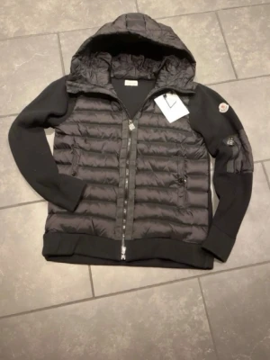 Moncler cardigan - Snygg svart cardigan från Moncler med quiltad front och rygg, stickade ärmar och huva. Cardigan har dragkedja framtill, två sidofickor med dragkedja och Moncler-logga på ärmen. Perfekt för kalla vinterdagar och riktigt trendig stil.