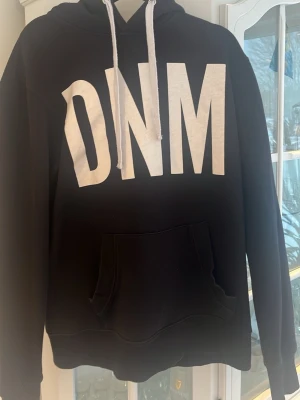 Svart hoodie från Denim Day & Co. - Svart hoodie från Denim Day & Co. med stor vit DNM-logga framtill. Klassisk känguruficka och vita dragsnören i huvan. Tillverkad i mjukt bomullsmaterial, perfekt för en avslappnad streetwear-look.