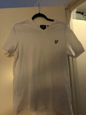 Vit t-shirt med v-ringning från Lyle & Scott - Vit t-shirt från Lyle & Scott med v-ringad hals och korta ärmar. På bröstet sitter den klassiska gula loggan med en broderad fågel. Tillverkad i mjuk bomull för en skön känsla. Enkel och stilren design som passar till det mesta.