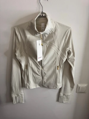 Vit träningsjacka från Lululemon storlek 6 - Snygg vit träningsjacka från Lululemon med hög krage och guldfärgade dragkedjor. Jackan har figurnära passform, långa ärmar med tumhål och två fickor framtill. Tillverkad i ett stretchigt funktionsmaterial som passar perfekt till träning.