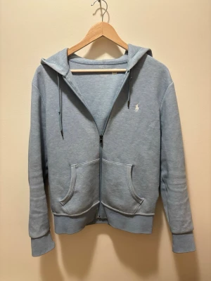 Ljusblå hoodie från Polo Ralph Lauren - Snygg ljusblå hoodie med dragkedja från Polo Ralph Lauren. Klassisk design med huva och dragsnören, två stora fickor framtill och den ikoniska vita logon broderad på bröstet. Tillverkad i mjuk bomullsblandning för skön komfort.