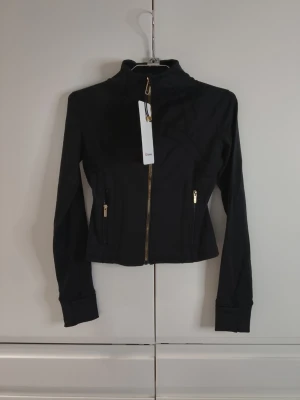 Svart träningsjacka från Lululemon Size 2 - Snygg svart träningsjacka från Lululemon med guldiga dragkedjor och hög krage. Jackan har figurnära passform, långa ärmar och två fickor framtill. Perfekt för gymmet eller löprundan. Materialet är stretchigt och andas bra.