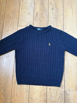 Mörkblå kabelstickad tröja Polo Ralph Lauren - Kabelstickad mörkblå tröja från Polo Ralph Lauren med gul broderad logga på bröstet. Tröjan är i mycket bra skick och tyget är mjukt och fräscht. Tröjan är något små i storleken. Hör gärna av er vid frågor. 