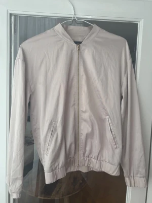 Ljusbeige bomberjacka från Soaked - Snygg ljusbeige- rosa bomberjacka från Soaked med dragkedja framtill och två snedställda fickor. Jackan har klassisk bombermodell med ribbad nederkant och är tillverkad i ett tunt, mjukt material. Perfekt för en stilren och avslappnad look.