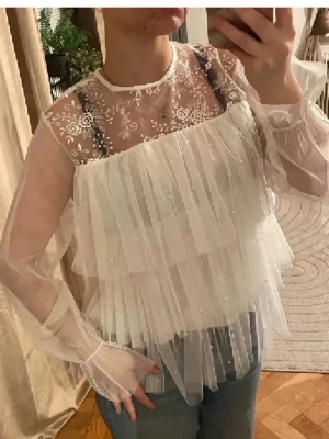 Vit volangblus med spetsdetaljer - Säljer en superfin vit blus med transparent mesh, broderade blommor upptill och flera lager volanger framtill. Blusen har långärmade ärmar i tunn tyll och rund halsringning. Perfekt för dig som gillar romantisk och trendig stil. By Malina liknade