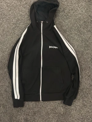 Svart hoodie från Palm Angels - Snygg svart hoodie från Palm Angels med vit dragkedja och vita ränder längs ärmarna. Klassisk huva och fickor framtill. Märkets logga broderad på bröstet. Perfekt för en streetwear-look. Tillverkad i mjukt material för extra komfort.