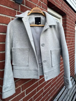Ljusgrå ulljacka från Lager  - Stilren ljusgrå ulljacka från Lager med rak passform, stora framfickor och klassisk krage. Jackan har tre knappar framtill och detaljerade manschetter. Perfekt för dig som gillar minimalistisk och modern stil.