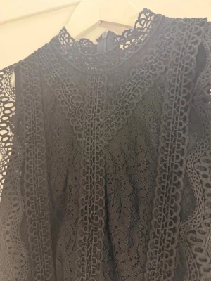 Svart spetsblus med hög krage - Säljer en svart blus i spets med hög krage och ärmlös design. Blusen har fina spetsdetaljer längs kanterna och ett elegant mönster över hela plagget. Perfekt att styla med jeans eller kjol för en trendig look.
