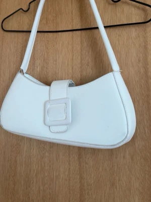 Vit axelremsväska med spänne - Snygg vit handväska i avlång modell med axelrem och dekorativt spänne framtill. Väskan har minimalistisk design och är tillverkad i slätt konstläder. Perfekt accessoar för att lyfta din outfit.