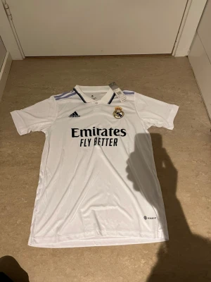Vit Real Madrid matchtröja Adidas - Snygg vit matchtröja från Real Madrid med klubbmärke och Adidas-logga på bröstet. Tröjan har svarta detaljer vid kragen och lila ränder på axlarna. Tillverkad i lätt och ventilerande material, perfekt för fotboll eller träning.
