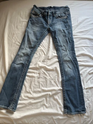 Blå bootcut jeans med broderade fickor - Snygga blå bootcut jeans med slitningar och broderade detaljer på bakfickorna. Jeansen har klassisk femficksdesign och kontrastsömmar. Perfekta för en avslappnad och trendig look.