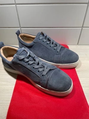 Blå mockasneakers från Louboutin - Snygga blå sneakers i mocka från Christian Louboutin med klassisk låg siluett och ikonisk röd sula. Skorna har matchande blå skosnören och diskreta sömdetaljer. Perfekta för dig som vill sticka ut med exklusiv design och lyxig känsla.