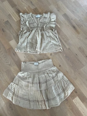 Beige set från Neo Noir med volanger, stl 36 - Säljer ett superfint beige set från Neo Noir med volangdetaljer. Toppen har korta ärmar och v-ringning med rysch, kjolen är kort med smock i midjan och flera lager volanger. Perfekt för en trendig och avslappnad look.