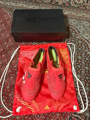 Röda Adidas Predator fotbollsskor - Säljer ett par röda Adidas Predator fotbollsskor med svarta och vita detaljer. Skorna har snörning och är designade för bra grepp och kontroll på planen. Perfekta för dig som vill sticka ut på fotbollsplanen.