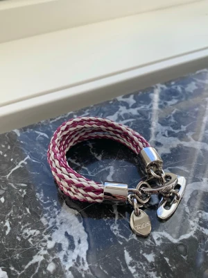 Flätat armband från Tod's - Snyggt armband från Tod's med flätad design i vinrött och vitt. Armbandet har silverfärgade metalldetaljer och en rund bricka med Tod's-loggan. Stängs med ett robust metallspänne. Perfekt accessoar för att lyfta din stil. Made in Italy 🇮🇹