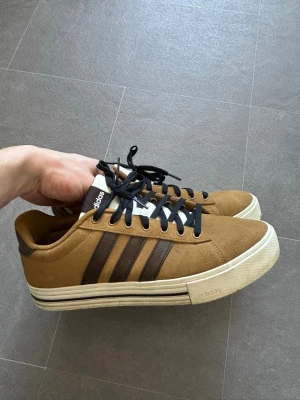 Bruna Adidas sneakers med mocka - Snygga bruna sneakers från Adidas med klassiska tre mörkbruna ränder på sidan. Skorna har svart snörning, vit tunga och vit sula med bruna detaljer. Ovandelen är i mocka och modellen är låg, perfekt för en avslappnad och cool stil.