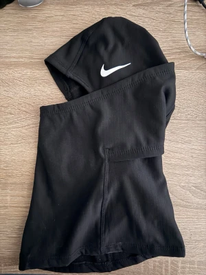 Svart balaclava från Nike - Svart balaclava från Nike med vit logga på sidan. Tillverkad i ett stretchigt och mjukt material som sitter tajt runt huvudet och halsen. Perfekt för kalla dagar eller sportaktiviteter utomhus.