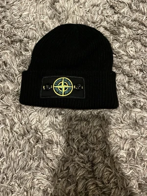 Svart ribbstickad mössa Stone Island - Säljer en svart ribbstickad mössa från Stone Island med den klassiska kompass-loggan framtill på en patch. Mössan har uppvikt kant och är tillverkad i ett mjukt, värmande material som passar perfekt för kalla dagar.