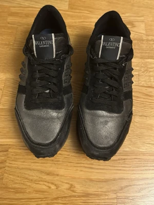 Svarta sneakers från Valentino Garavani - Snygga svarta sneakers från Valentino Garavani med coola nitar på sidorna och detaljer i både mocka och läder. Skorna har svarta skosnören och en robust sula som ger en sportig look. Perfekta för dig som gillar exklusiva och trendiga sneakers.