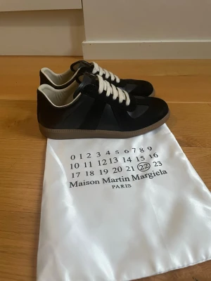 Maison Margiela svarta sneakers - Svarta sneakers från Maison Martin Margiela med vita snören och beige sula. Klassisk low-top modell med stilren design och detaljer i mocka och läder. Perfekta för dig som gillar minimalistisk och trendig streetstyle.