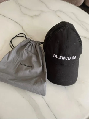 Svart keps från Balenciaga - Snygg svart keps från Balenciaga med vit broderad logga framtill. Klassisk böjd skärm och justerbar rem baktill. Kepsen är i bomull och kommer med original dustbag och tag. Perfekt accessoar för en trendig streetstyle-look.