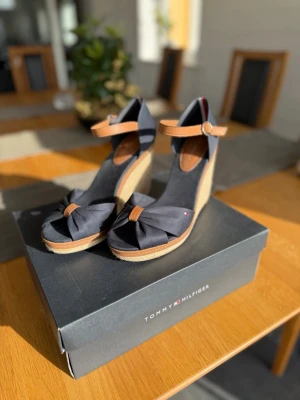 Kilklackar från Tommy Hilfiger - Snygga kilklackar från Tommy Hilfiger i marinblå textil med bruna detaljer och öppen tå. Skorna har en flätad kilklack i naturfärgad jute och justerbart ankelband med spänne. 