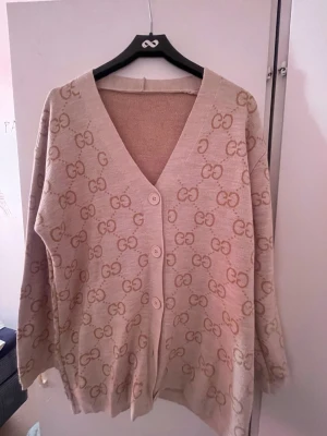 Beige cardigan med GG-mönster  - Snygg beige kofta med klassiskt GG-mönster i ljusbrunt över hela plagget. Koftan har v-ringning, tre knappar framtill och långa ärmar. Perfekt för dig som vill ha en lyxig och trendig look.