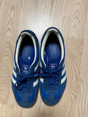 Blå Adidas Originals sneakers striped  - Snygga blå sneakers från Adidas Originals med klassiska vita ränder på sidorna och gummisula med mönster. Skorna har låg profil och är tillverkade i mocka och textil med vita detaljer runt öppningen. Perfekt för dig som gillar retrostil.