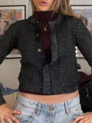 Svart glittrig kofta med knappar - Snygg svart glittrig kofta med strukturstickat mönster och dekorativa knappar framtill. Kortare modell med ribbade muddar och satinliknande kant vid knäppningen. Perfekt att styla över en topp eller polo. Storlek xs