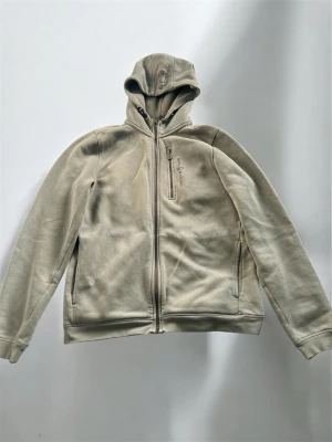 Ljus grön hoodie med dragkedja och fickor - Snygg beige hoodie från Peak Performance med dragkedja framtill, huva och två sidofickor. Bröstficka med dragkedja och broderad logga på bröstet. Tillverkad i mjukt material som passar perfekt till en avslappnad stil.