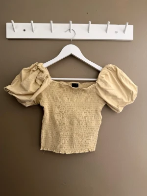Beige puffärm topp från Gina Tricot - Trendig beige topp från Gina Tricot med smockad kropp och stora puffärmar. Croppad modell med fyrkantig halsringning, perfekt för en cool och avslappnad stil. Tillverkad i mjukt material som känns skönt mot huden.
