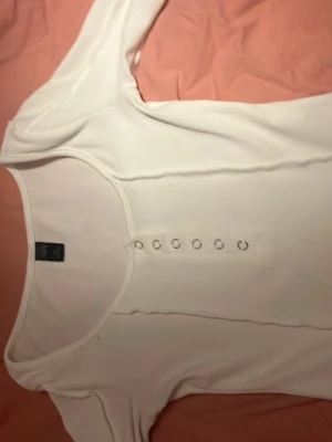 Vit ribbad långärmad topp  - Snygg vit långärmad topp med ribbad struktur och dekorativa knappar framtill. Toppen har en rundad halsringning och sitter figurnära för en clean och enkel look. Perfekt att styla med jeans eller kjol.