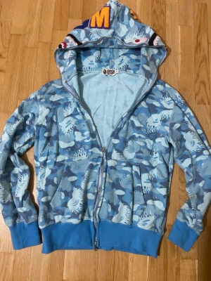 Blå camo hoodie från BAPE - Säljer en blå camo hoodie från BAPE med dragkedja och ikoniskt hajmotiv på huvan. Hoodien har ribbade muddar, fickor fram och färgglada broderade detaljer på huvan. Tillverkad i mjuk bomullsblandning, perfekt för streetwear-stilen. Den är XL men passar egentligen XS. 160-170 längd.