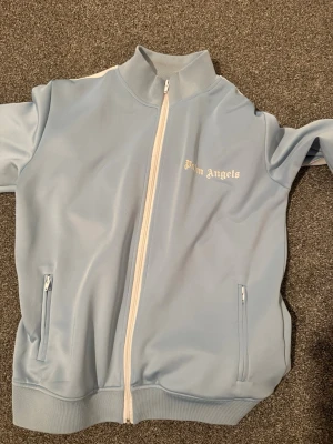 Ljusblå tracksuit från Palm Angels - Snygg ljusblå tracksuit  från Palm Angels med vit dragkedja och vita detaljer på axlarna. Jackan har hög krage, två fickor med dragkedja och logga tryckt på bröstet. Tillverkad i ett glansigt material som ger en sportig vibe. Byxorna har lite tråd försvunnit som ni kan se på bilden men annars är dom helt perfekta. 