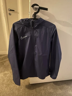 Mörkblå Nike vindjacka med huva - Snygg mörkblå vindjacka från Nike med dragkedja framtill och huva. Jackan har en vit Nike-logga på bröstet och är tillverkad i ett lätt, vindtätt material. Perfekt för blåsiga dagar och sportiga outfits. Endast använd 2 gånger! Den är som ny bara att prislappen inte är kvar. Priset går att diskutera!