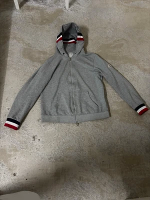 Moncler cardigan - Snygg grå hoodie från Moncler med dragkedja framtill och huva. Detaljer i rött, vitt och blått på huva och ärmslut ger en sportig vibe. Moncler-logga på ärmen. Tillverkad i mjuk bomullsblandning, perfekt för chill dagar.