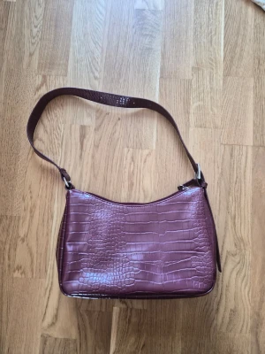 Vinröd axelväska i crocopräglat skinn - Säljer en vinröd axelväska med crocopräglat mönster och justerbar axelrem. Väskan har en stilren, rektangulär form och är tillverkad i glansigt konstläder. Perfekt för dig som vill ha en trendig accessoar med lite edge.