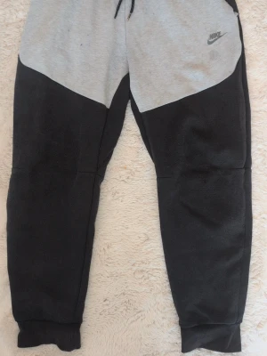 Nike mjukisbyxor svart/grå - Nike mjukisbyxor i svart och ljusgrått med logga på vänster sida. Byxorna har resår i midjan med dragsko, muddar vid bensluten och sidofickor. Tillverkade i mjukt sweatshirtmaterial som är perfekt för chill eller träning. 