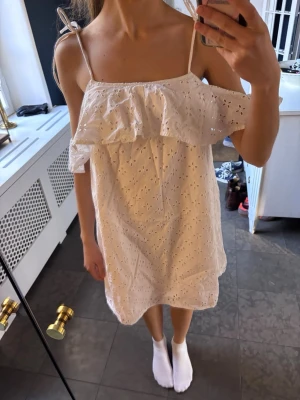 Vit offshoulder klänning med volang - Supersöt vit kortklänning i bomull med broderade detaljer och volang upptill. Klänningen har offshoulder-design med smala axelband som knyts och öppen rygg. 
