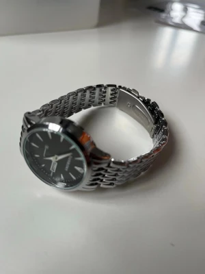 Silverfärgad klocka - Stilren klocka med svart urtavla och datumvisning. Armbandet är i blankt silverfärgat metall och har ett klassiskt länkmönster. Perfekt accessoar för dig som gillar en clean och tidlös look. Inte jättestor så passar dig med mindre handleder