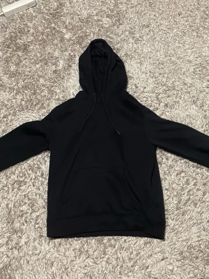Mörkblå basic hoodie med huva - En stilren mörk blå hoodie med huva och dragsnören. Hoodien har en klassisk känguruficka framtill och ribbade muddar vid ärmslut och nederkant. Perfekt för en avslappnad look och enkel att matcha med andra plagg.