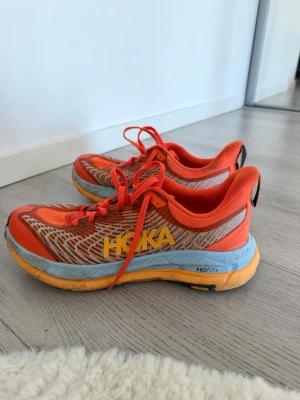 Orange Hoka sneakers med blå sula - Snygga sneakers från Hoka i en riktigt fräsch orange färg med gula detaljer och blå sula. Ovandelen är i mesh med grå och vita mönster för extra ventilation. Perfekta för dig som gillar sportig stil och vill sticka ut med färg.