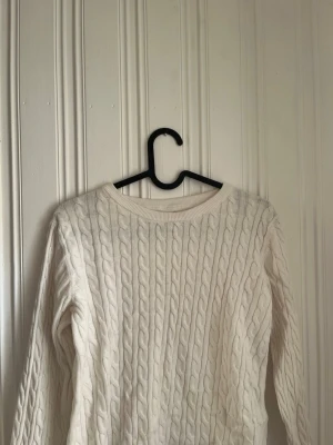 Vit kabelstickad tröja - Kabelstickad tröja i mjuk vit bomullsblandning med rund halsringning och långa ärmar. Klassisk design med ribbade muddar vid hals, ärmslut och nederkant. Perfekt för lager på lager och enkel att matcha med jeans eller kjol.