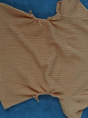 Beige croppad topp med rynk - Trendig croppad topp i beige med rynkad struktur och korta ärmar. Toppen har en rundad halsringning med knytband och elastisk kant som ger en cool och avslappnad look. Perfekt till höga jeans eller kjol.