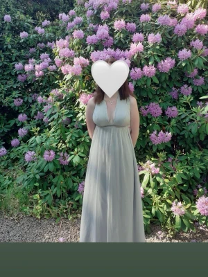 Ljusgrå långklänning med v-ringning - Elegant ljusgrå långklänning med djup v-ringning och breda axelband. Klänningen har ett flytande fall och markerad midja, perfekt för sommarens speciella tillfällen. Materialet känns lätt och svalt, vilket gör den bekväm att bära under varma dagar.