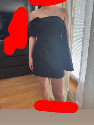 Svart oneshoulder klänning - Trendig svart oneshoulder klänning med asymmetrisk design och draperad detalj över axeln. Klänningen har en stilren och modern look, perfekt för dig som vill sticka ut. Materialet känns slätt och har en lätt glans.