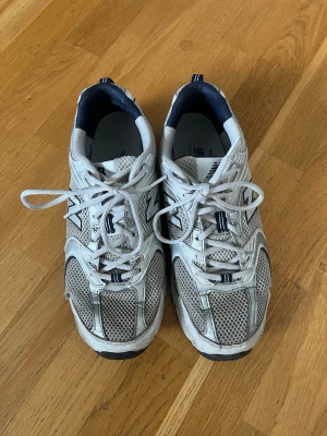 Vita New Balance 530 sneakers - Säljer ett par klassiska New Balance 530 sneakers i vitt med marinblå och silverdetaljer. Skorna har meshpaneler för extra ventilation, robust sula och ikoniska N-loggan på sidan. Perfekta för dig som gillar retrostil och sportig vibe.