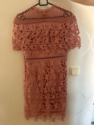 Rosa spetsklänning med blommönster - Supersöt rosa kortklänning med blommig spets och hög hals. Klänningen har korta ärmar och fina volangdetaljer. Perfekt för sommarens alla tillfällen! Materialet känns som spets och polyester.