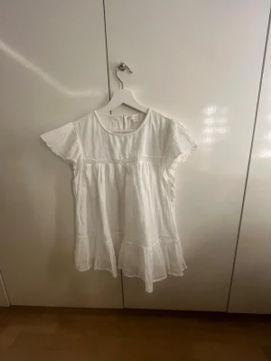 Vit luftig klänning med volanger - Söt vit klänning i ett luftigt och lätt tyg med volangdetaljer nedtill och korta ärmar. Klänningen har rund halsringning och avslappnad passform, perfekt för sommardagar. Materialet ser ut att vara bomull eller linne för en skön känsla.