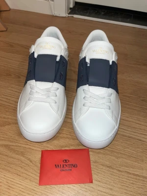 Valentino Garavani opens vita/mörkblå - Snygga vita sneakers från Valentino Garavani med bred mörkblå rem över vristen och vita skosnören. Skorna har en stilren design i läder och subtila nitar på hälen. Perfekta för dig som vill ha en exklusiv och modern look.